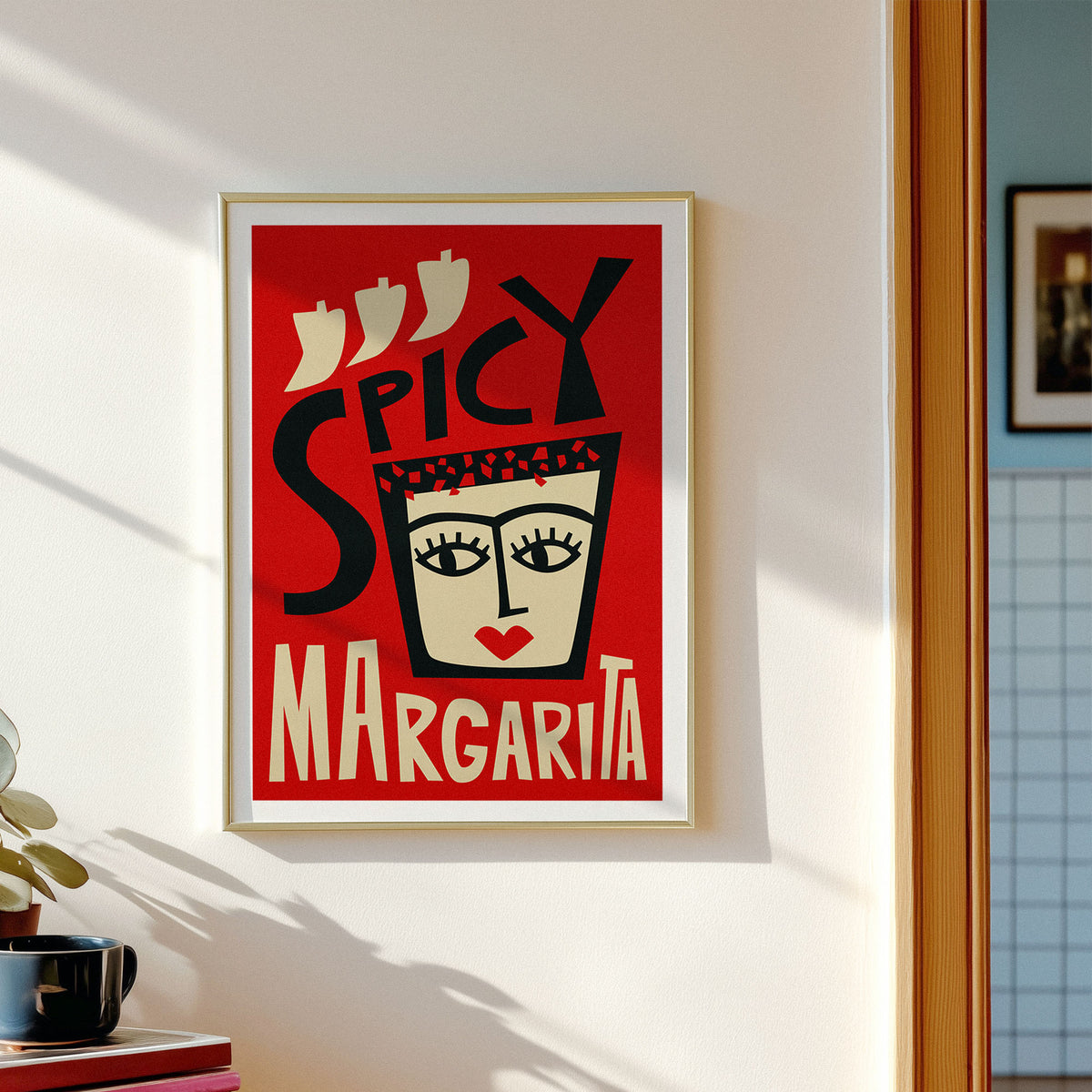 A framed art print featuring a retro-style illustration of a margarita glass with the words 'SPICY MARGARITA' on a red background, hanging on a light gray wall in a dining room setting with wooden chairs and a table.
