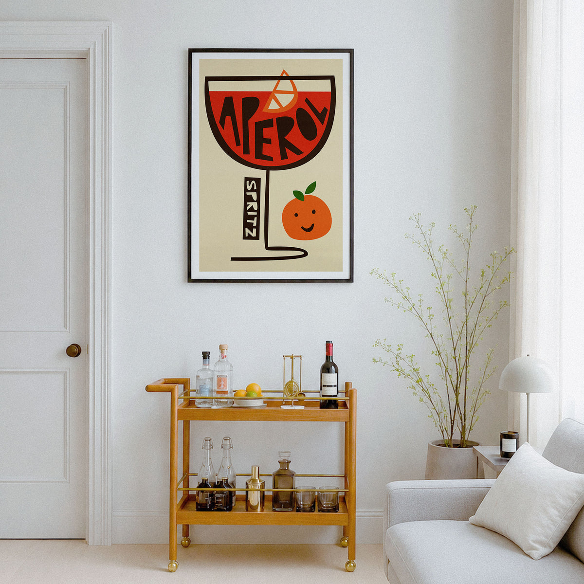 A framed art print featuring a retro-style orange cocktail glass with the word 'SPRITZ' and a smiling orange character, displayed on a white wall above a wooden bar cart in a modern living room setting.