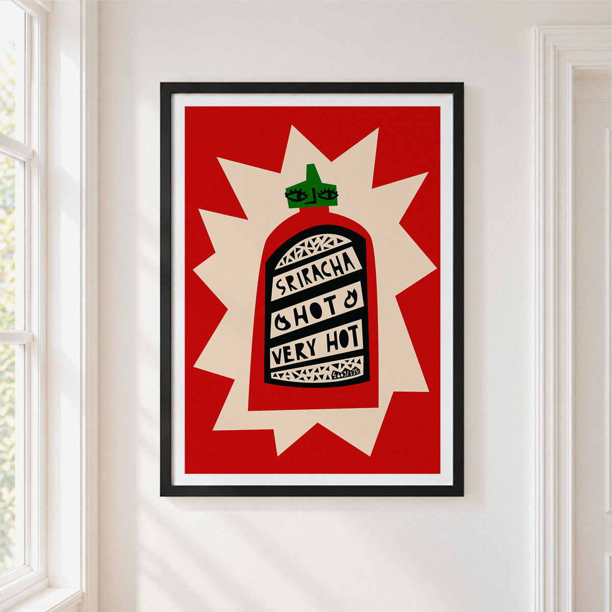 Framed wall art featuring a stylized hot sauce bottle with the words 'SRIRACHA HOT VERY HOT' on a red and cream background, hung on a white wall next to a window.