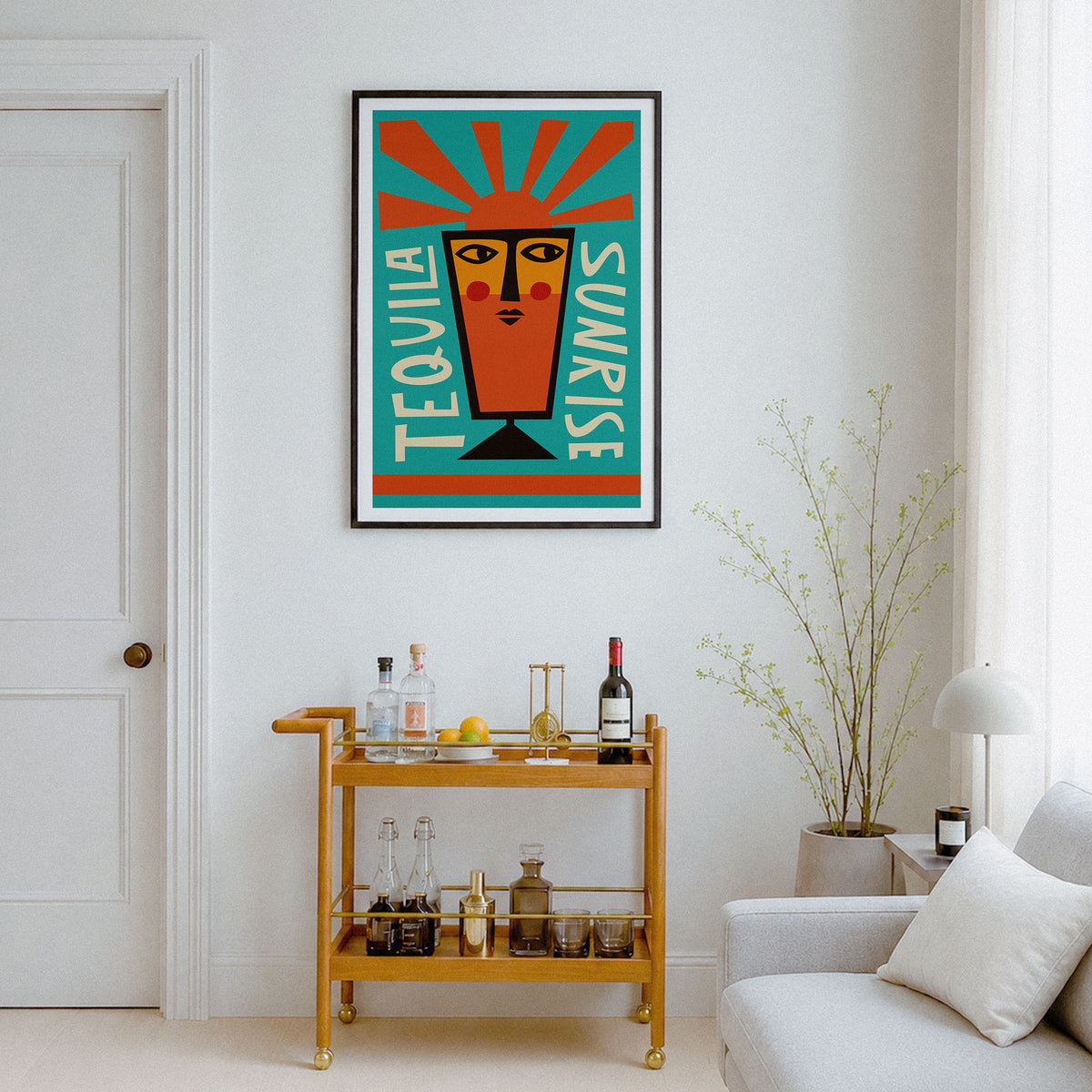 A framed art print featuring a stylized tequila glass with the words 'TEQUILA' and 'SUNRISE' in a retro geometric design, hanging on a white wall in a modern living room setting above a wooden bar cart.