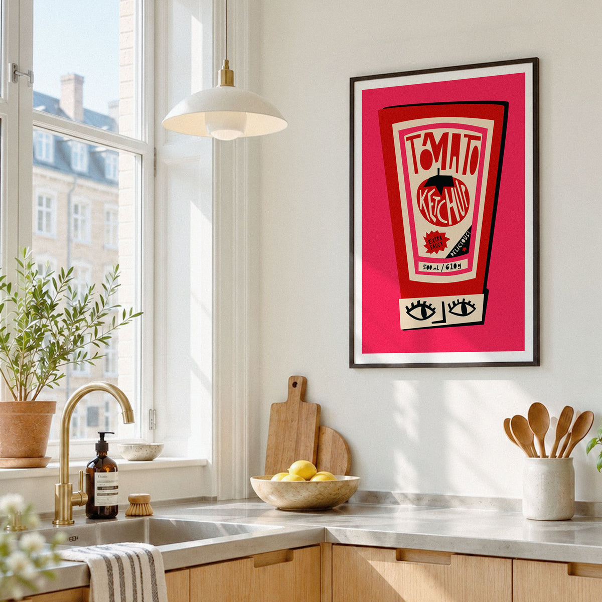 A framed art print featuring a retro red and pink tomato ketchup can graphic hangs on a white kitchen wall above a marble countertop with wooden cabinets, a gold faucet, and kitchen accessories.