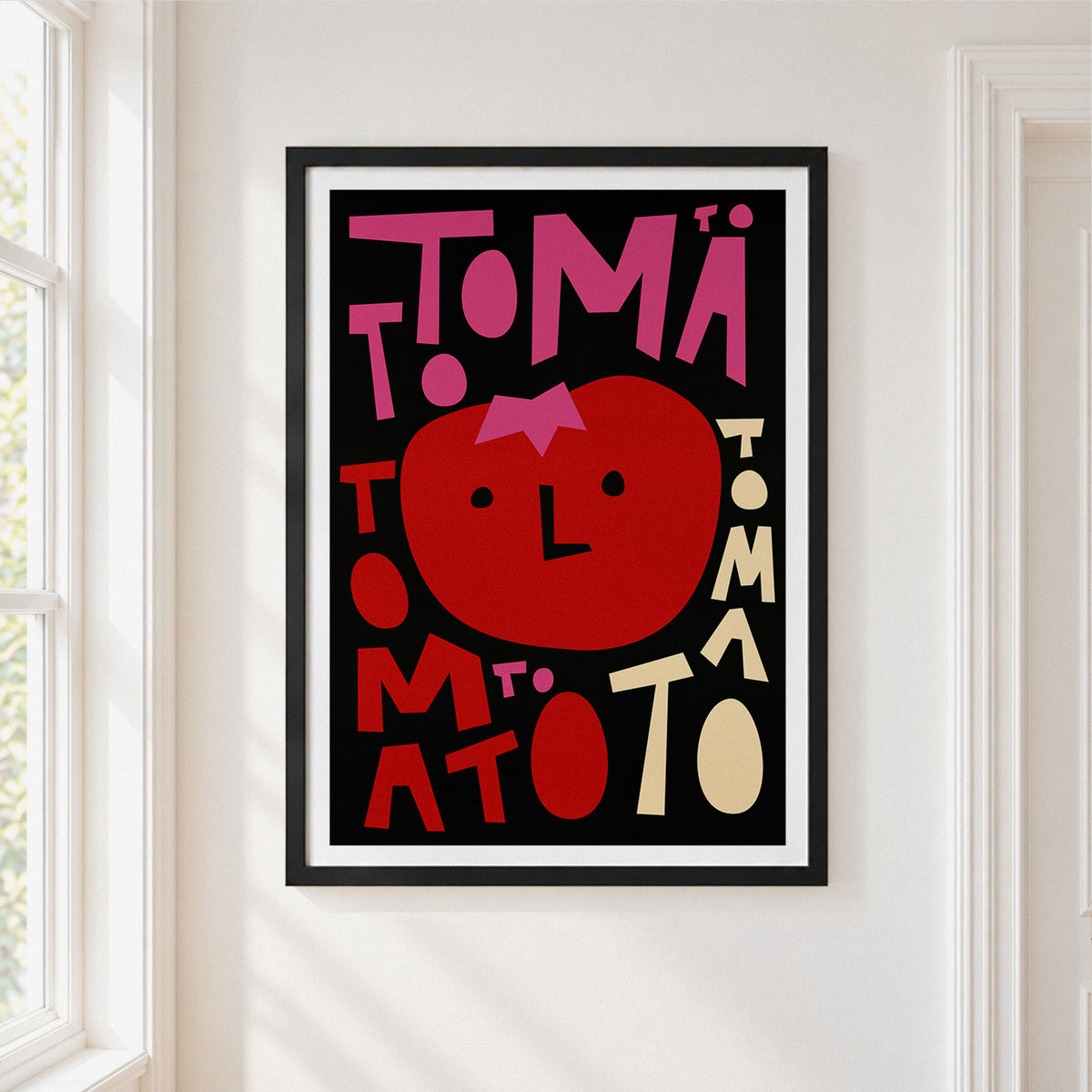 A framed graphic art print featuring a stylized red tomato with a pink bow and the word 'TOMATO' in bold pink and red lettering on a black background, hung on a white wall next to a window.
