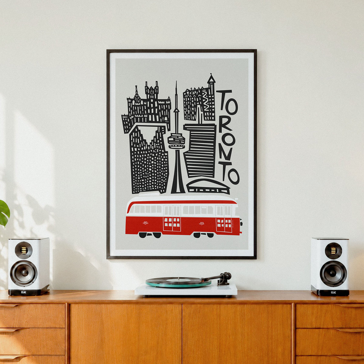 A framed Toronto cityscape art print hangs on a white wall above a wooden credenza, flanked by two white speakers and a vinyl record player. The print features black silhouettes of Toronto landmarks and a red transit bus on a light gray background.