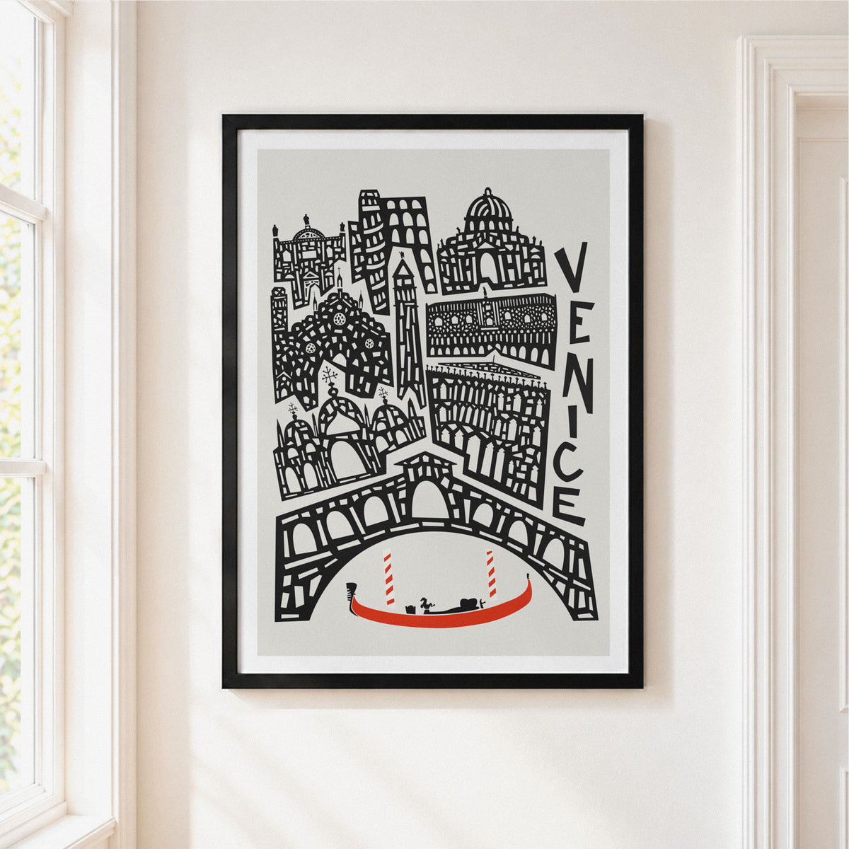 A framed art print featuring a black and white hand-drawn illustration of Venice with a small red gondola, hanging on a light beige wall next to a window.