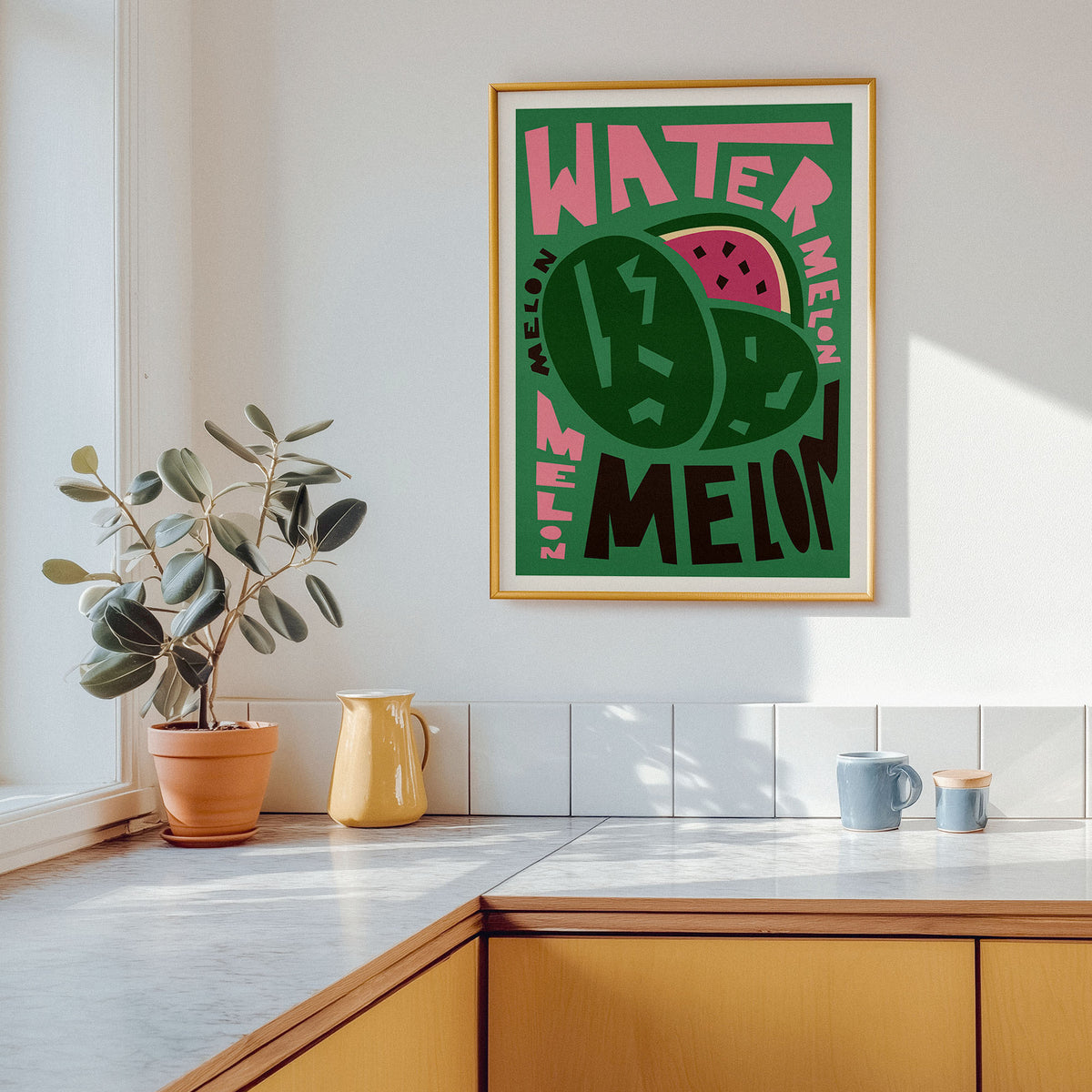 A framed watermelon-themed art print hangs on a white kitchen wall above a countertop with a potted plant, yellow pitcher, and blue mugs. The print features bold pink and black text reading 'WATER MELON MELON' with green watermelon illustrations.