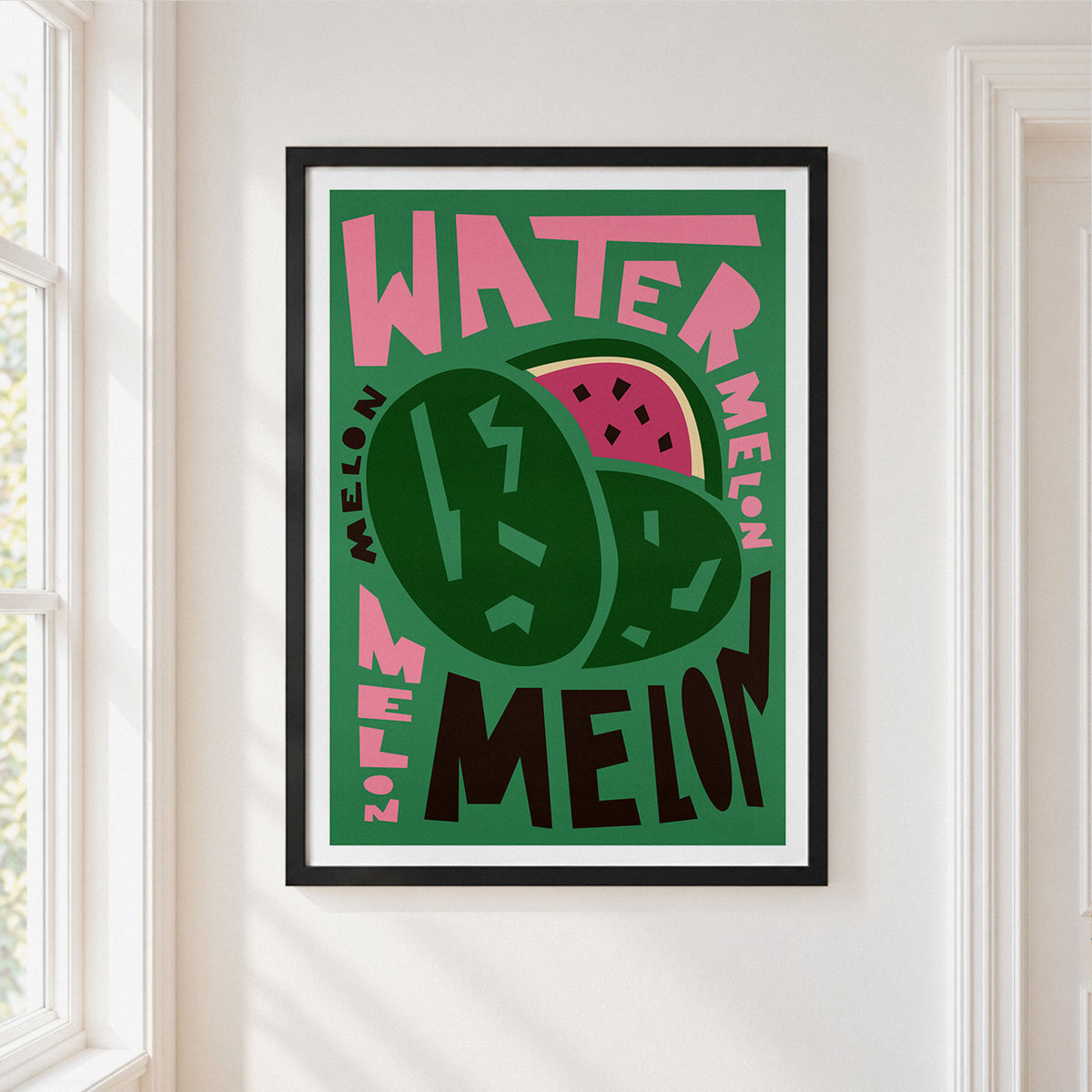 A framed art print featuring a stylized watermelon illustration with the words 'WATERMELON' in bold pink and dark green lettering, hung on a white wall next to a window.