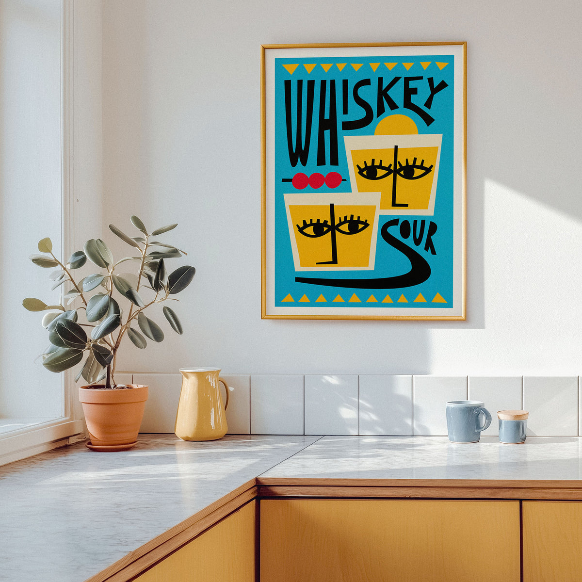 A framed art print featuring a retro graphic design with the words 'Whiskey Sour' and stylized cocktail glasses with eyes, hanging on a white kitchen wall above a counter with a potted plant and mugs.