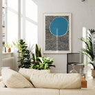 A large framed abstract blue and black print displayed in a modern white living room filled with plants, lending boldness and calm to the contemporary decor.