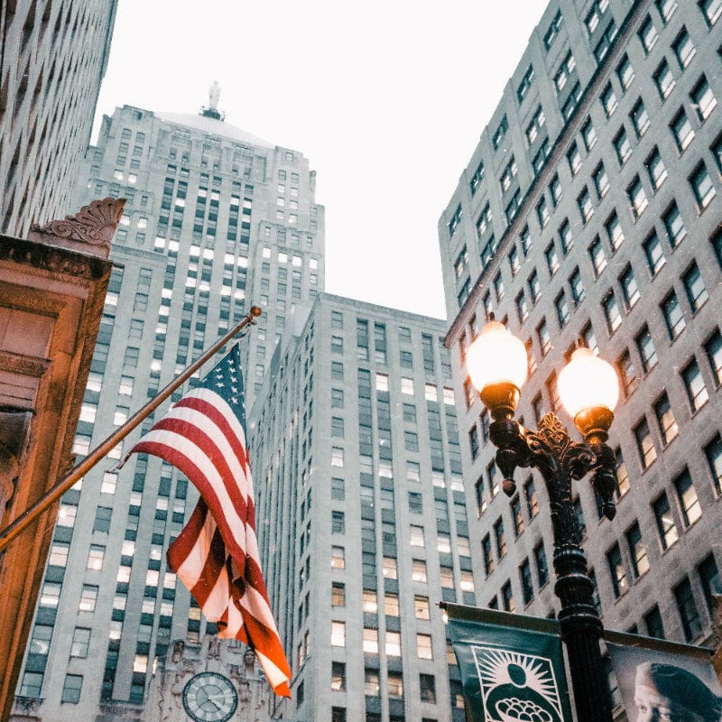The USA flag on a flag pole next to a lamp post below some tall skyscrapers.