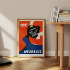 Aquarius print framed on the floor next to a pale wood desk and books in a clean, modern nook.