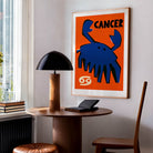 Cancer zodiac poster framed above a small table in a bright modern room with a lamp and warm wood tones.