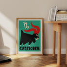 Capricorn print framed on the floor beside a pale wood desk with stacked books in a minimal home space.