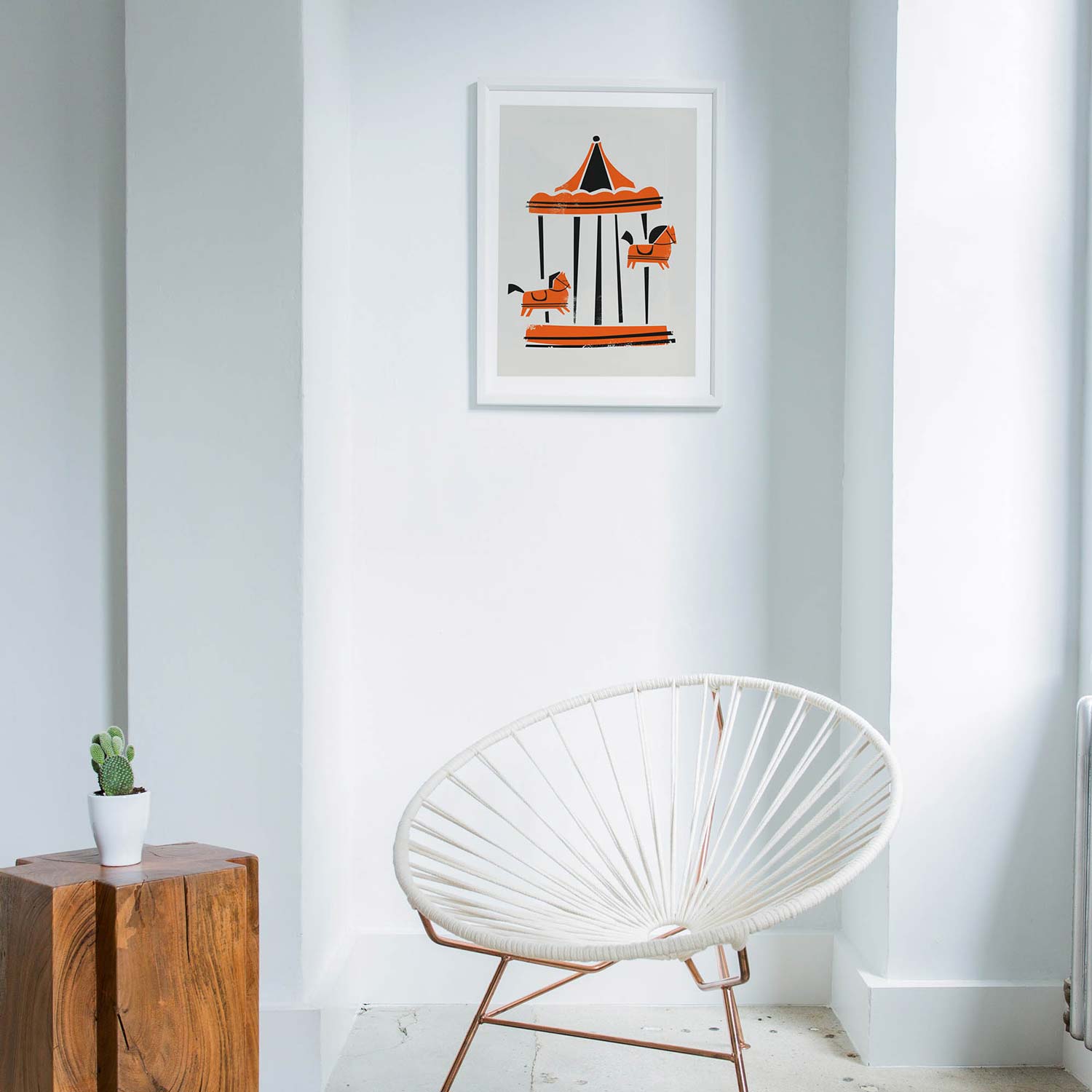 A framed art print of a whimsical carousel hanging on a white wall in a modern room with a white woven chair and a wooden side table.