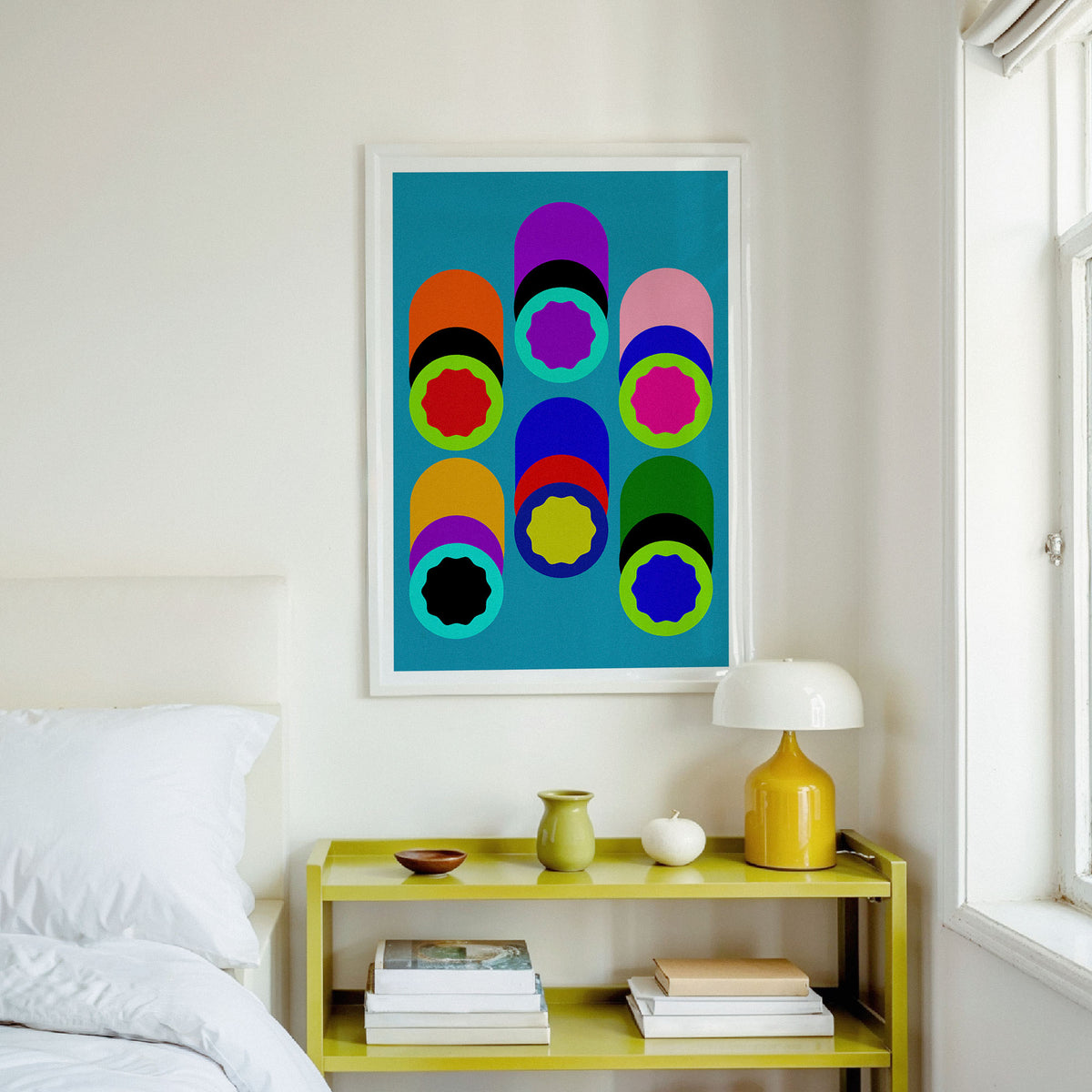 A framed abstract art print featuring colorful geometric circles on a teal background, hanging on a white wall above a yellow shelf in a bright bedroom setting.