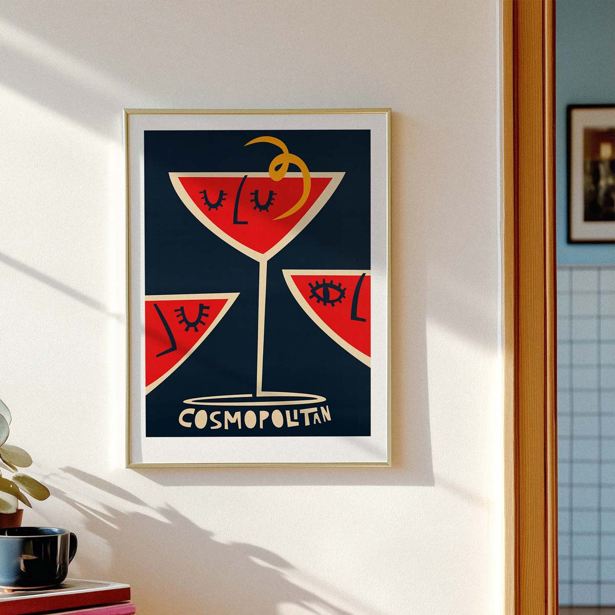 Framed wall art hanging on a white wall, featuring a stylized cocktail glass illustration in red, navy, cream, and gold with the word 'COSMOPOLITAN' at the bottom. Part of a kitchen scene is visible at the right edge.
