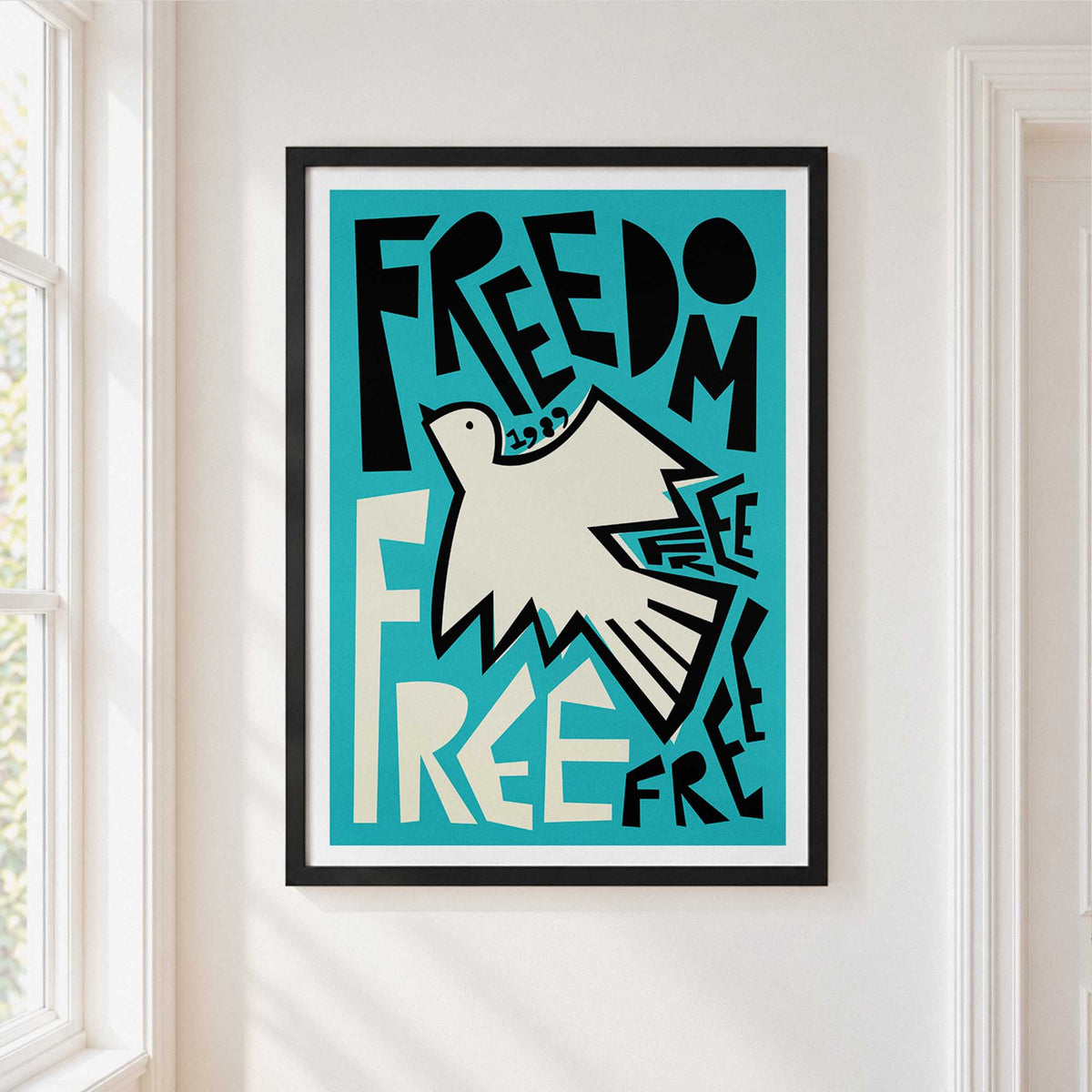A framed art print featuring a stylized white dove and the lettering typogrpahy 'FREEDOM' on a turquoise background, hanging on a white wall next to a window.
