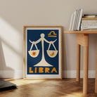 Libra print framed on the floor next to a wooden desk with stacked books and pale wall.