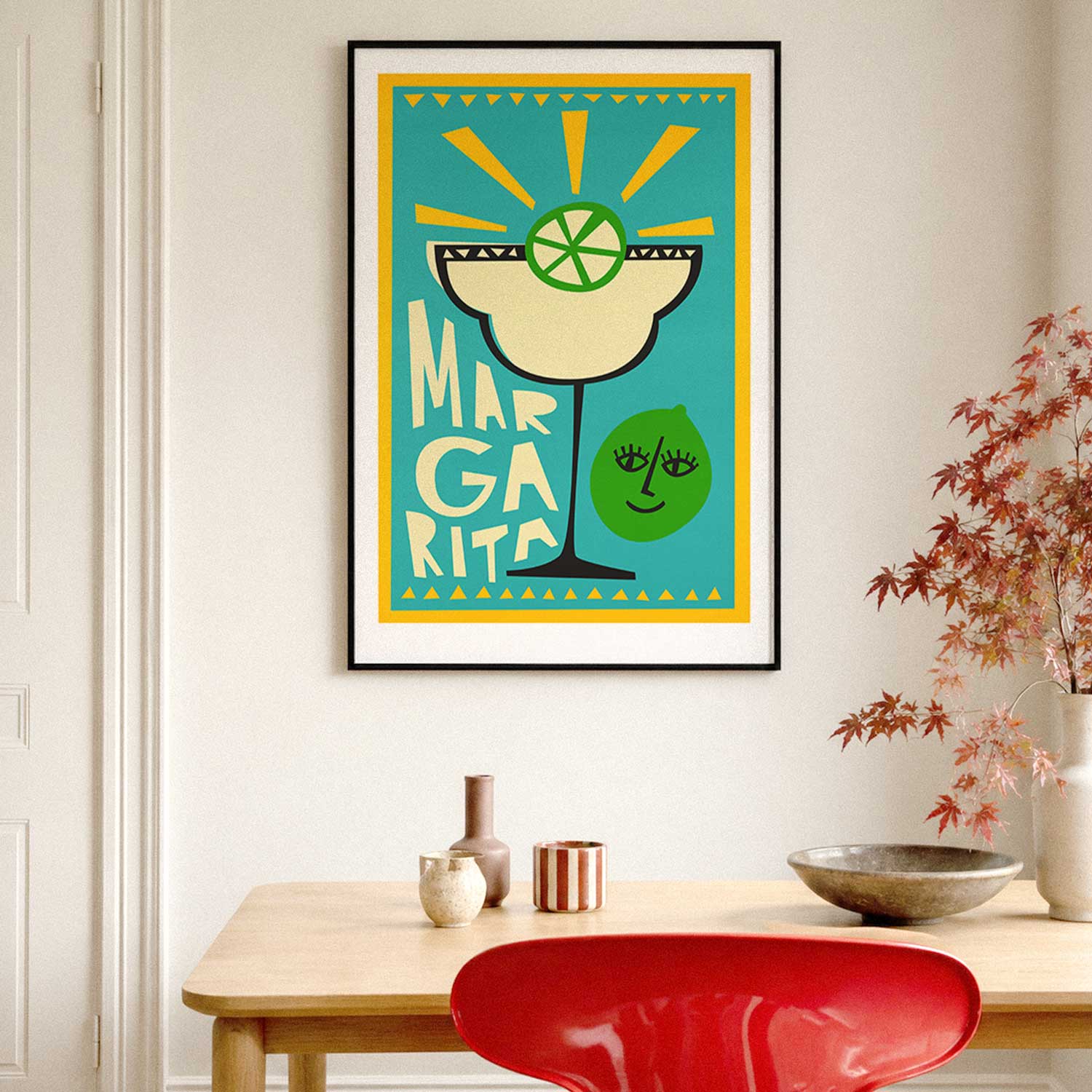 A framed art print featuring a colorful Margarita illustration hangs on a light beige wall above a wooden table with decorative vases, a bowl, and a red chair.