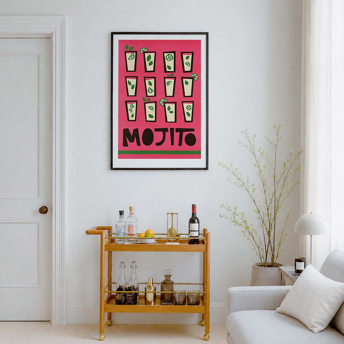 A framed art print featuring a retro pink background with green cocktail glasses and the word 'MOJITO' in bold black letters, displayed on a white wall in a modern living room setting above a wooden bar cart.