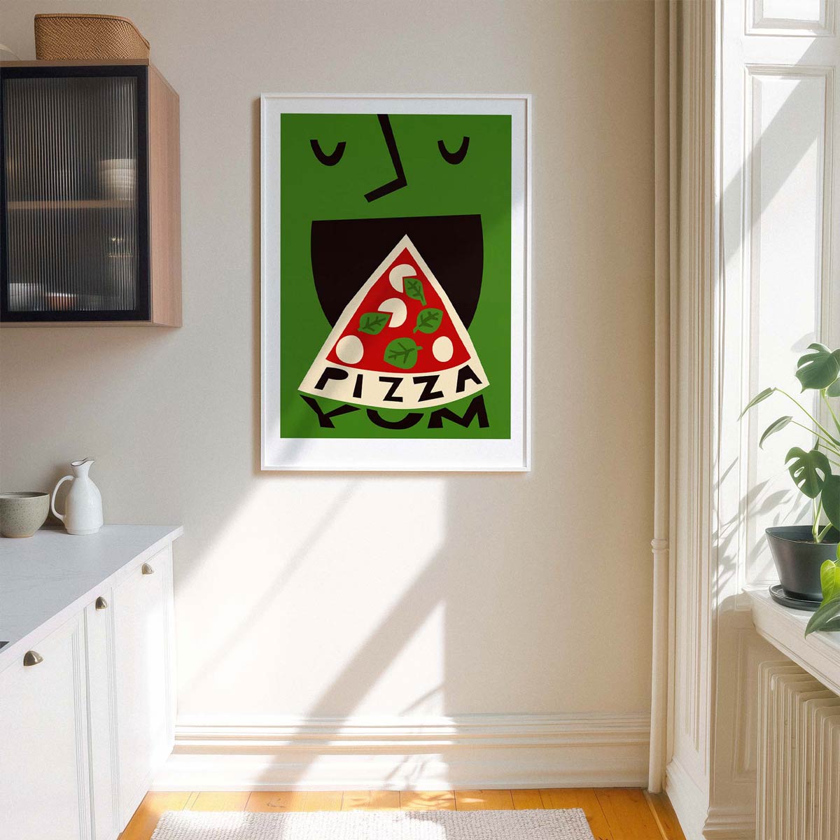 A framed art print featuring a whimsical illustration of a green face with a slice of pizza for a mouth and the word 'PIZZA' below it, displayed on a beige wall in a bright kitchen setting.