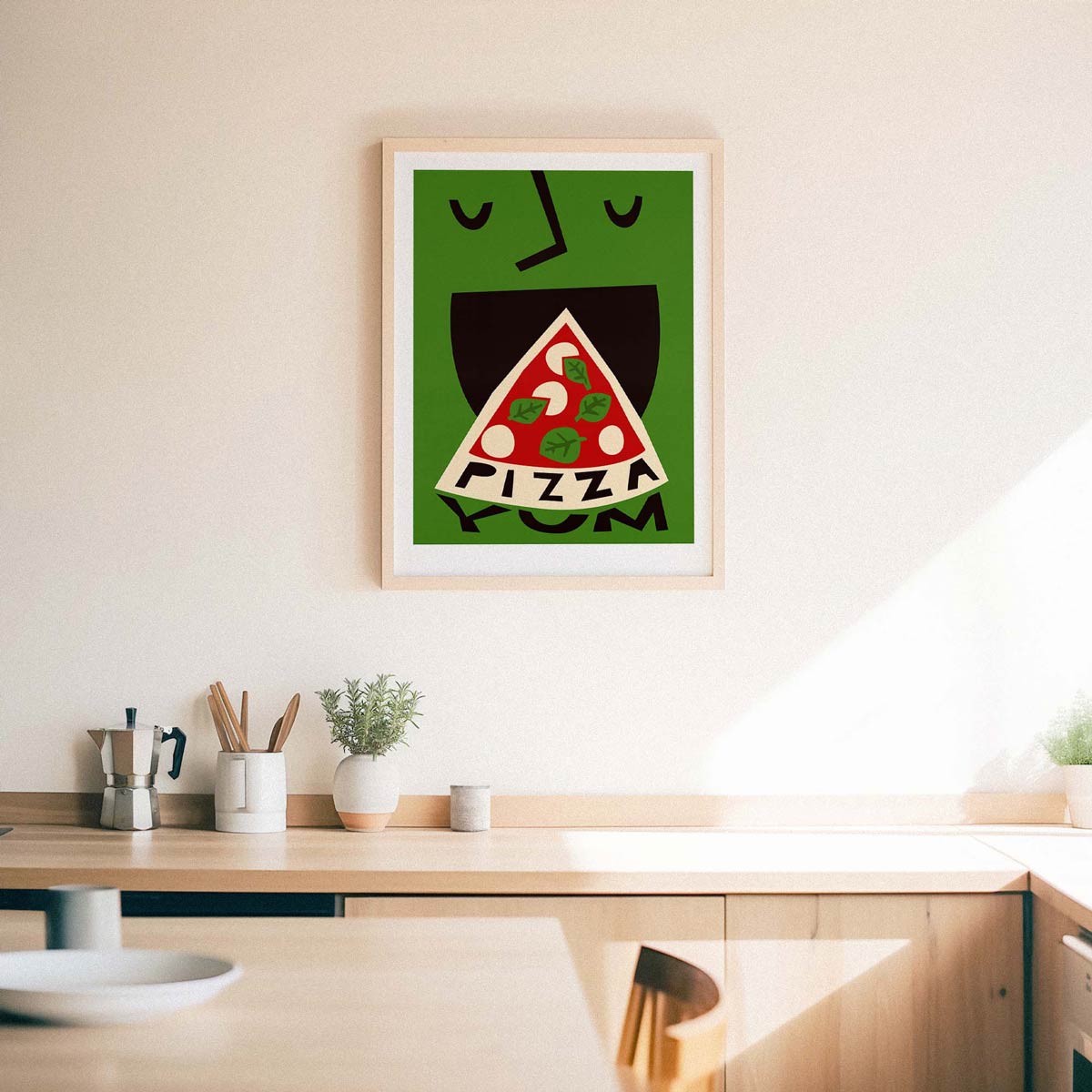 A framed art print featuring a stylized green face eating a slice of pizza with the word 'PIZZA' inside, hanging on a light beige wall above a wooden kitchen counter.