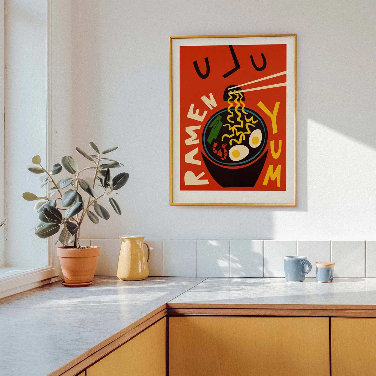A framed retro-style ramen poster hangs on a white kitchen wall above a countertop with a potted plant, yellow pitcher, and mugs. The poster features bold orange, red, black, and yellow artwork with the words 'RAMEN YUM'.