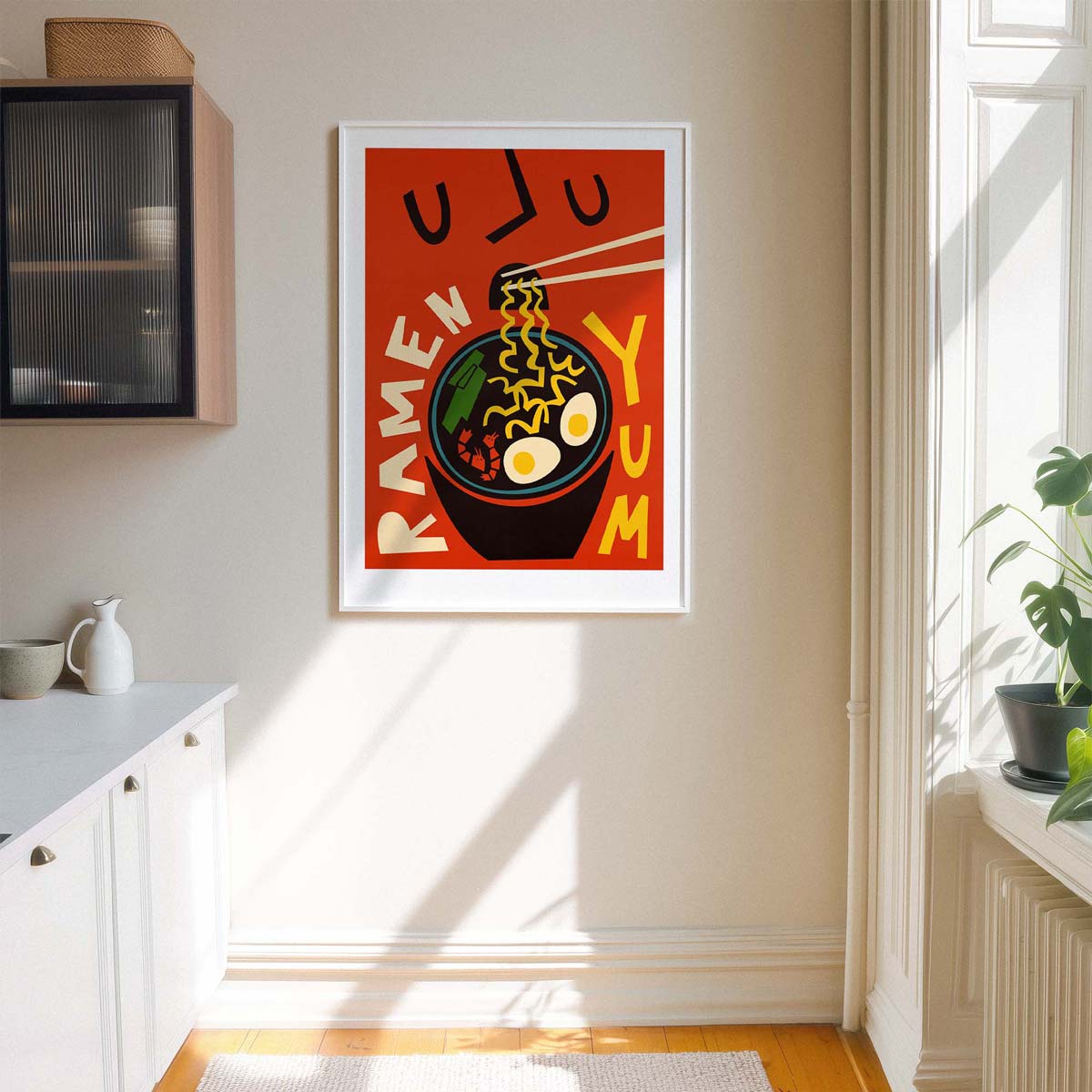 A framed vertical art print featuring a stylized illustration of a ramen bowl with chopsticks and the words 'RAMEN YUM' on a warm orange-red background, displayed on a beige wall in a sunlit kitchen interior.