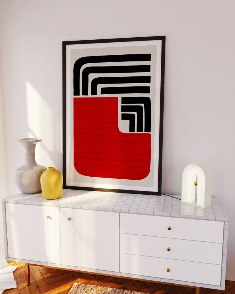 The Sol print framed and leaning against a white wall on a sideboard, with warm lighting that highlights its architectural shapes and striking modern palette.