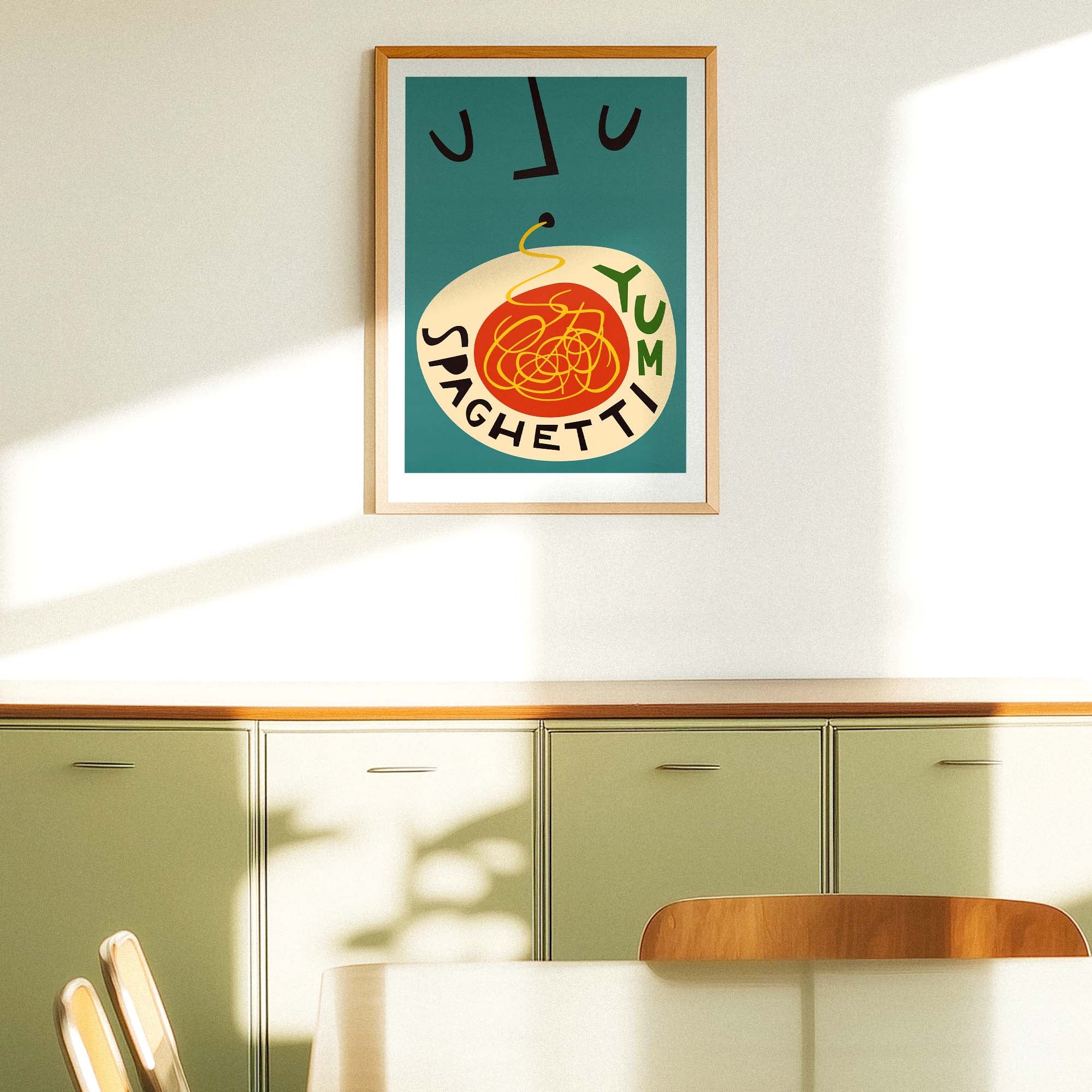 A framed art print featuring a retro illustration of spaghetti with the words 'YUM SPAGHETTI' on a teal background, hanging on a white wall above light green kitchen cabinets in a sunlit room.