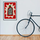 Sriracha hot sauce artwork framed on a clean white wall beside a bike and wooden floor.