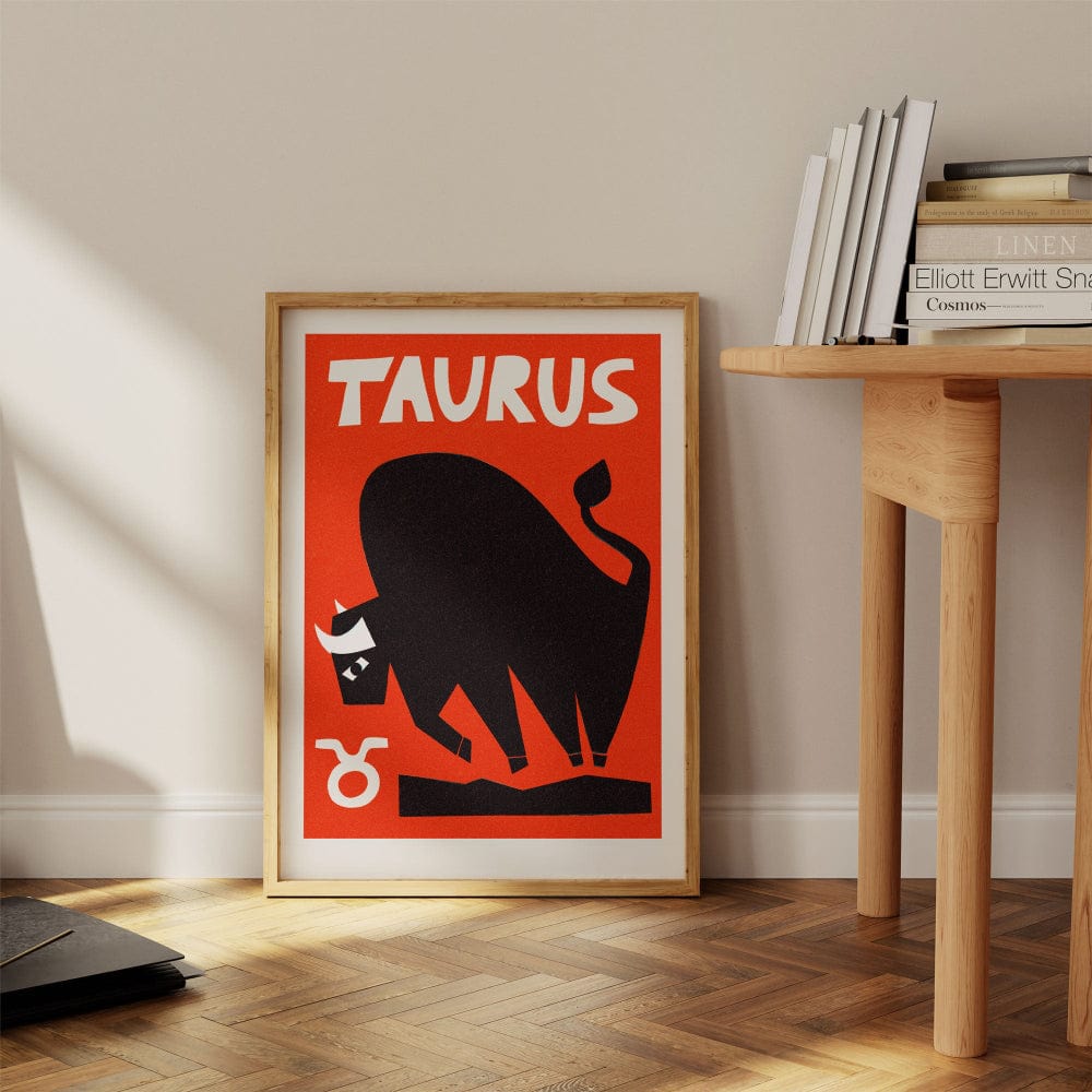 Taurus zodiac artwork framed on the floor beside a wood desk and books in a warm modern nook.