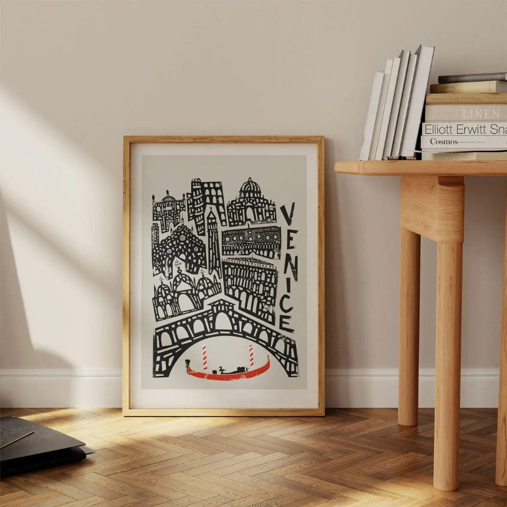 Framed Venice city print leaning against a wooden desk, showcasing its bold architectural lines and vibrant red gondola in a cosy, creative interior setting.