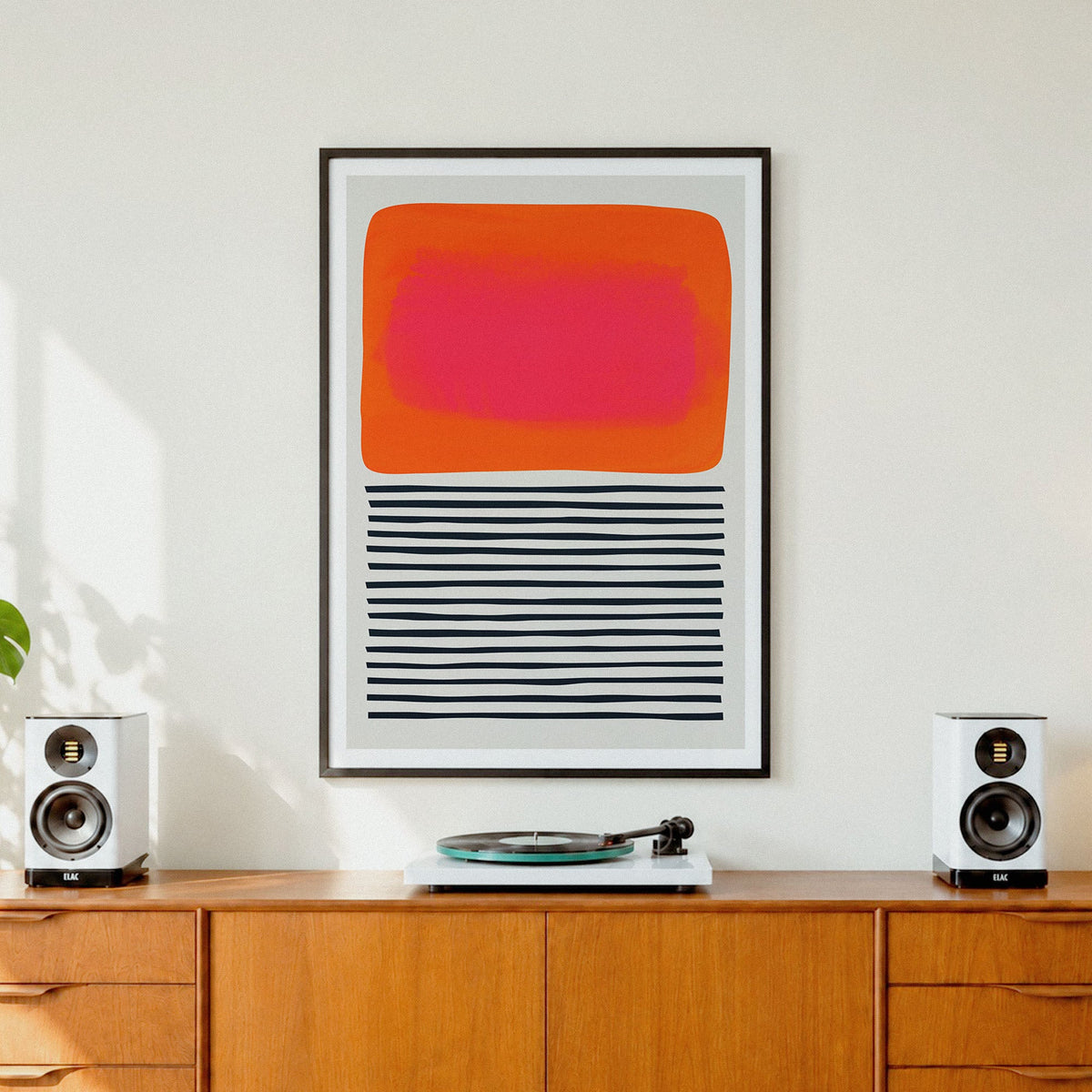 A framed abstract art print  'Sunset Ripples' featuring a bright orange and pink rectangle above black horizontal stripes, displayed on a wooden cabinet in a modern room setting with speakers and a record player.