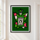 A framed art print featuring a stylized green Absinthe bottle with a jester hat, set against a dark green background with pink, white, and green abstract shapes. The print is displayed on a white wall next to a window.