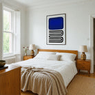 A modern bedroom featuring a large framed abstract art print with a bold blue and black geometric design on a white wall above a wooden bed. The room is styled with white bedding, beige textured throw blankets, matching wooden nightstands, and ceramic table lamps.