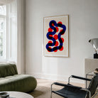 A framed abstract geometric art print with colorful twisting shape hangs on a light gray wall in a modern living room setting with a green sofa and black chair.