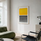 A framed abstract wall art print hangs on a light gray wall in a modern living room. The artwork features a large yellow rounded rectangle on the upper half and horizontal black stripes on the lower half, set in a thin light wood frame.