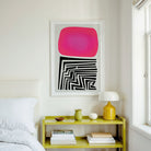 A framed abstract art print featuring a bright pink shape above black geometric lines, displayed on a white wall above a lime green nightstand in a bedroom setting.