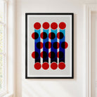 Framed abstract geometric wall art hanging on a white wall, featuring red and black circles over vertical blue and navy bars on a light gray field, with a black frame visible.