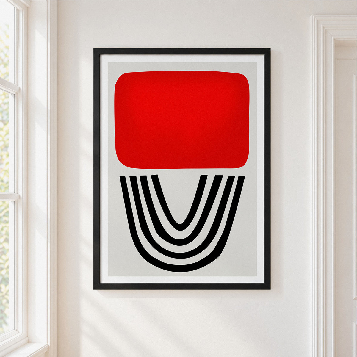 A framed abstract art print hanging on a white wall in a bright room. The print features a bold red rounded rectangle on a light gray background, with black curved lines below it, enclosed in a thin black frame.