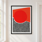 A framed abstract art print featuring a bold red circle, a red-orange geometric shape, and a black-and-white patterned lower section, displayed on a white wall next to a window.