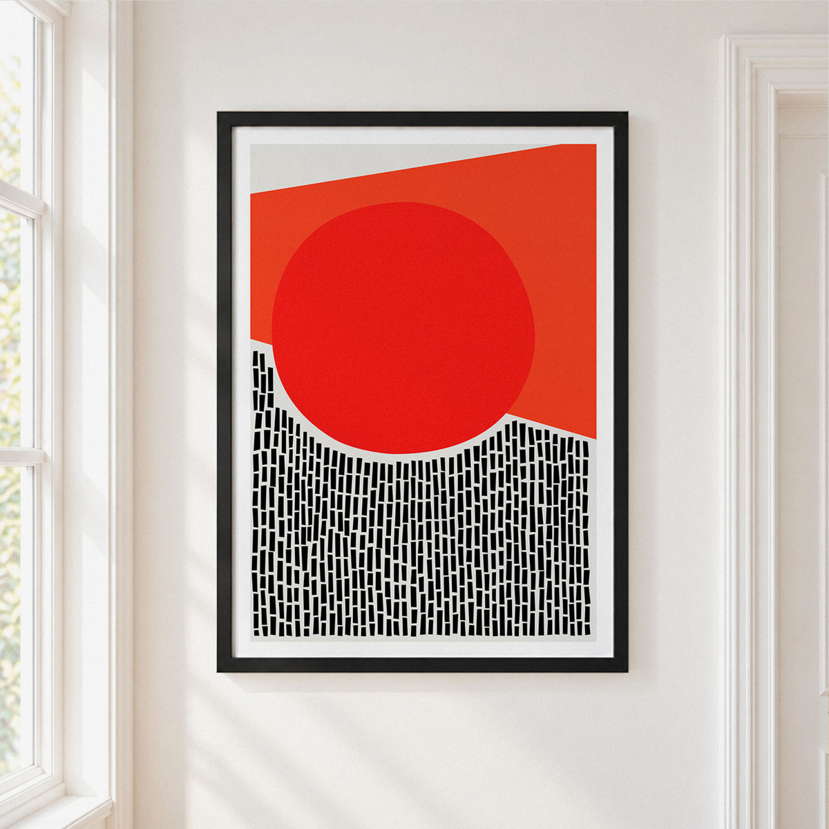 A framed abstract art print featuring a bold red circle, a red-orange geometric shape, and a black-and-white patterned lower section, displayed on a white wall next to a window.