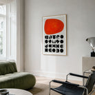 A framed abstract geometric art print with red shape and geometric black shapes underneath  shapes hangs on a light grey wall in a modern living room setting with a green sofa and black chair.