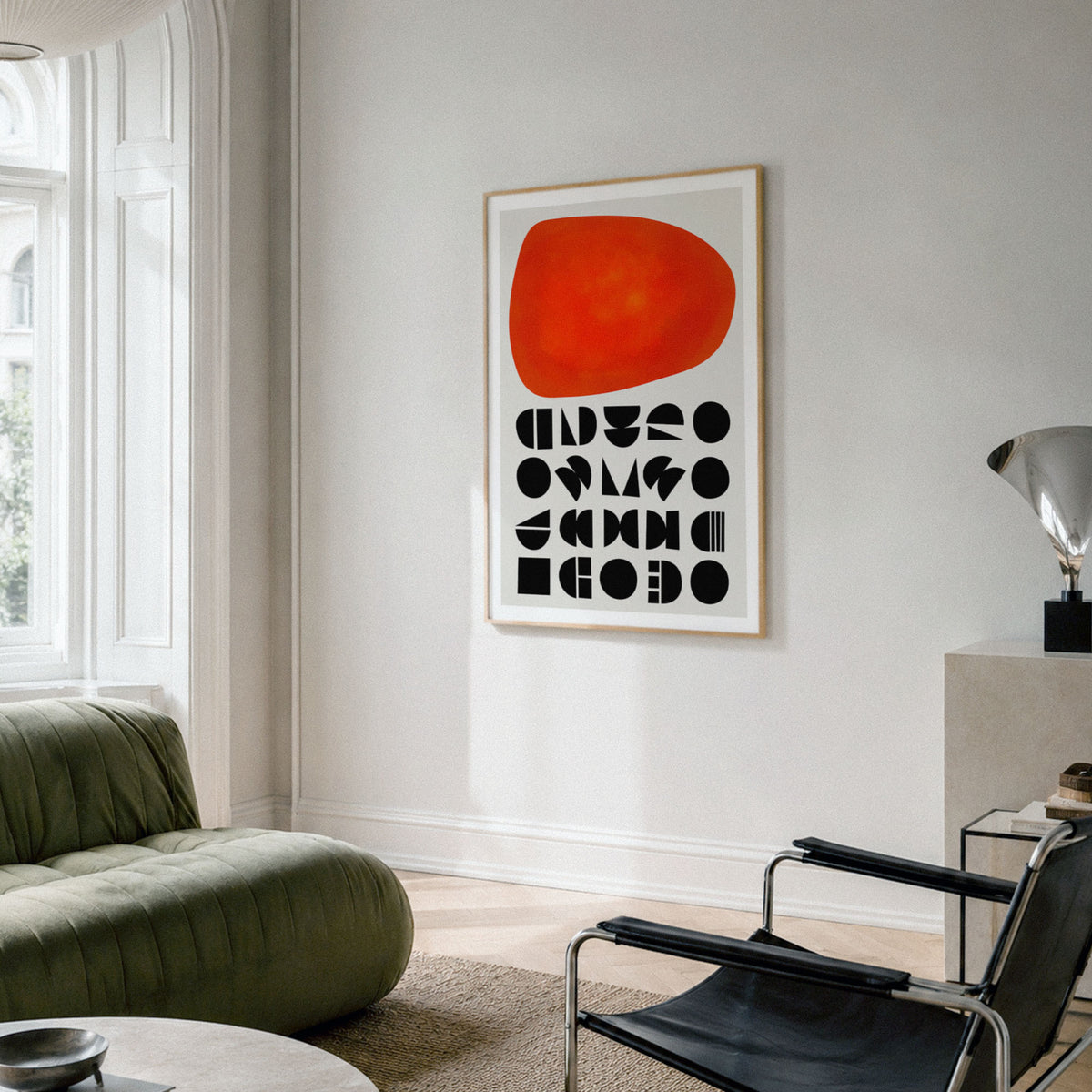 A framed abstract geometric art print with red shape and geometric black shapes underneath  shapes hangs on a light grey wall in a modern living room setting with a green sofa and black chair.