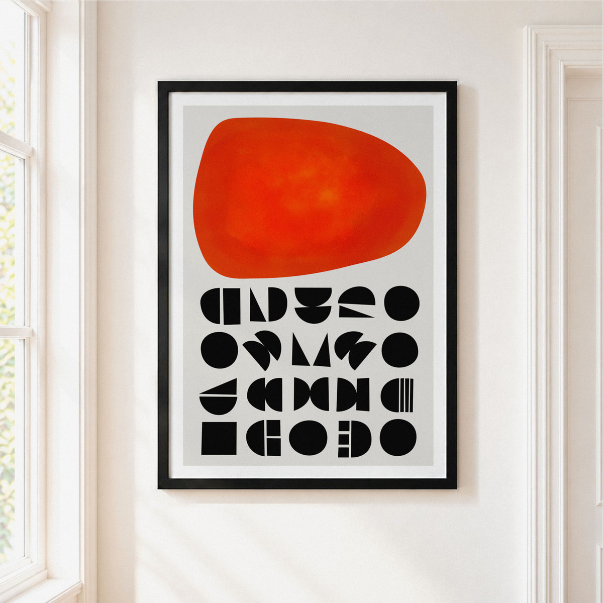Framed abstract wall art hanging on a white wall beside a window. The print features a large orange-red organic shape above rows of black geometric symbols on a light gray background, enclosed in a black frame.