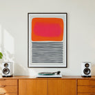 A framed abstract art print  'Sunset Ripples' featuring a bright orange and pink rectangle above black horizontal stripes, displayed on a wooden cabinet in a modern room setting with speakers and a record player.