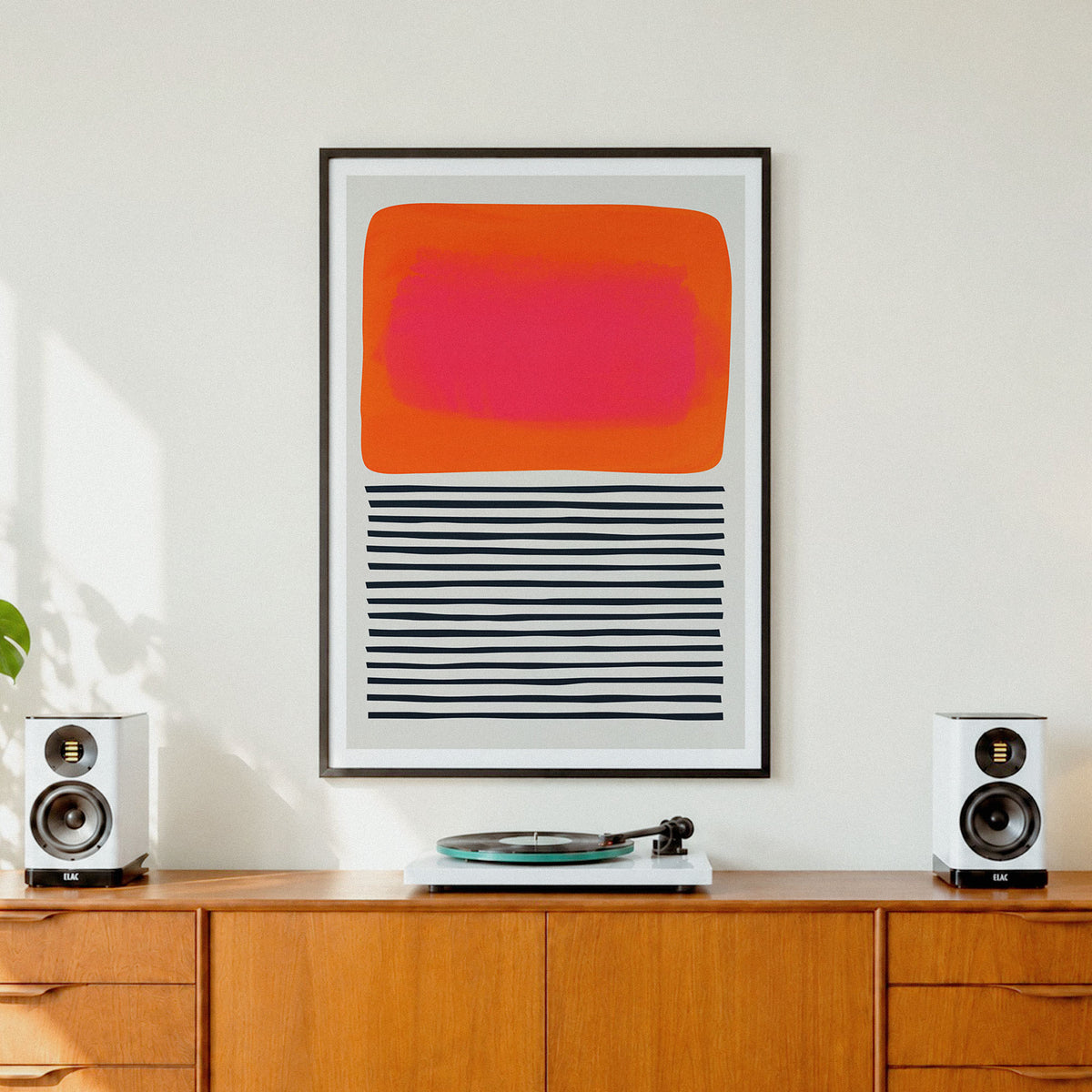 A framed abstract art print  'Sunset Ripples' featuring a bright orange and pink rectangle above black horizontal stripes, displayed on a wooden cabinet in a modern room setting with speakers and a record player.