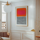 A framed abstract art print hangs on a white paneled wall in a bright, elegant room. The artwork features a vibrant pink and orange block on the top half and black horizontal stripes on the bottom half, set in a light wood frame.