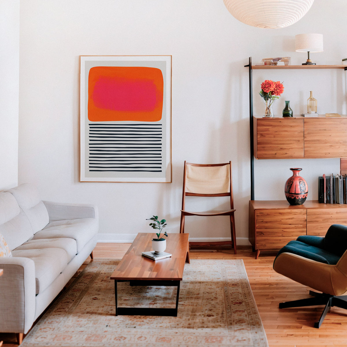 A modern living room featuring a framed abstract art print with a bright orange and pink rectangle and black horizontal stripes on a white background, hung on a white wall above a wooden coffee table.