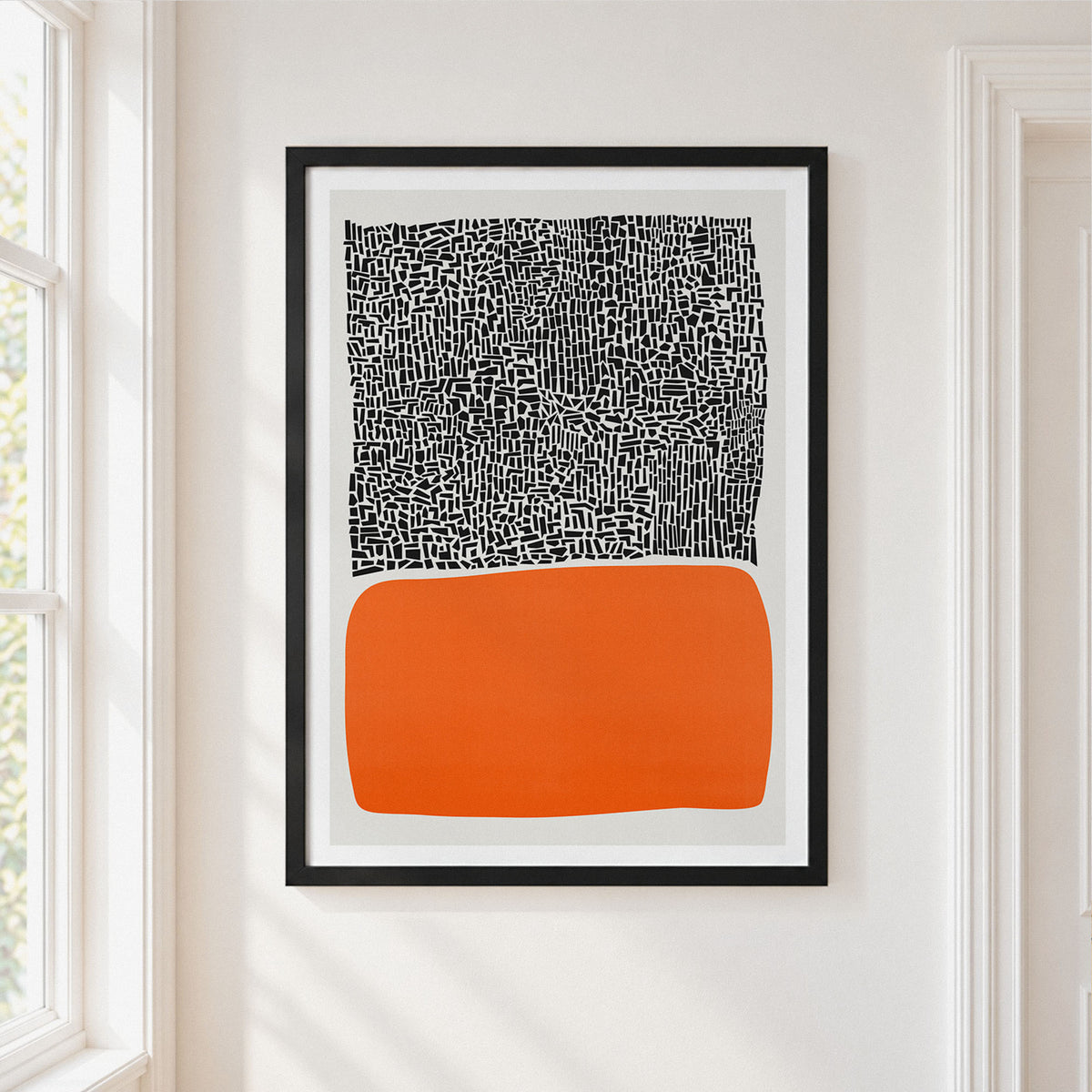Framed abstract wall art hanging on a white wall beside a window. The print has a black-and-white patterned upper section and a large orange lower section, enclosed in a black frame.